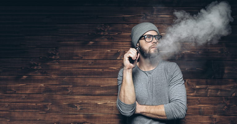 VAPING HIPSTER LEANING ON BICK WALL BLOWING SMOKE WEARING A GRAY CAP