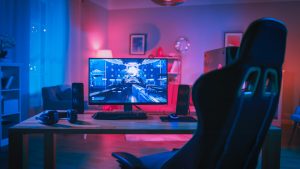 Personal Computer Gamer Rig with First-Person Shooter Game on Screen. Monitor Stands on the Table at Home. Cozy Room with Modern Design is Lit with Pink Neon Light.